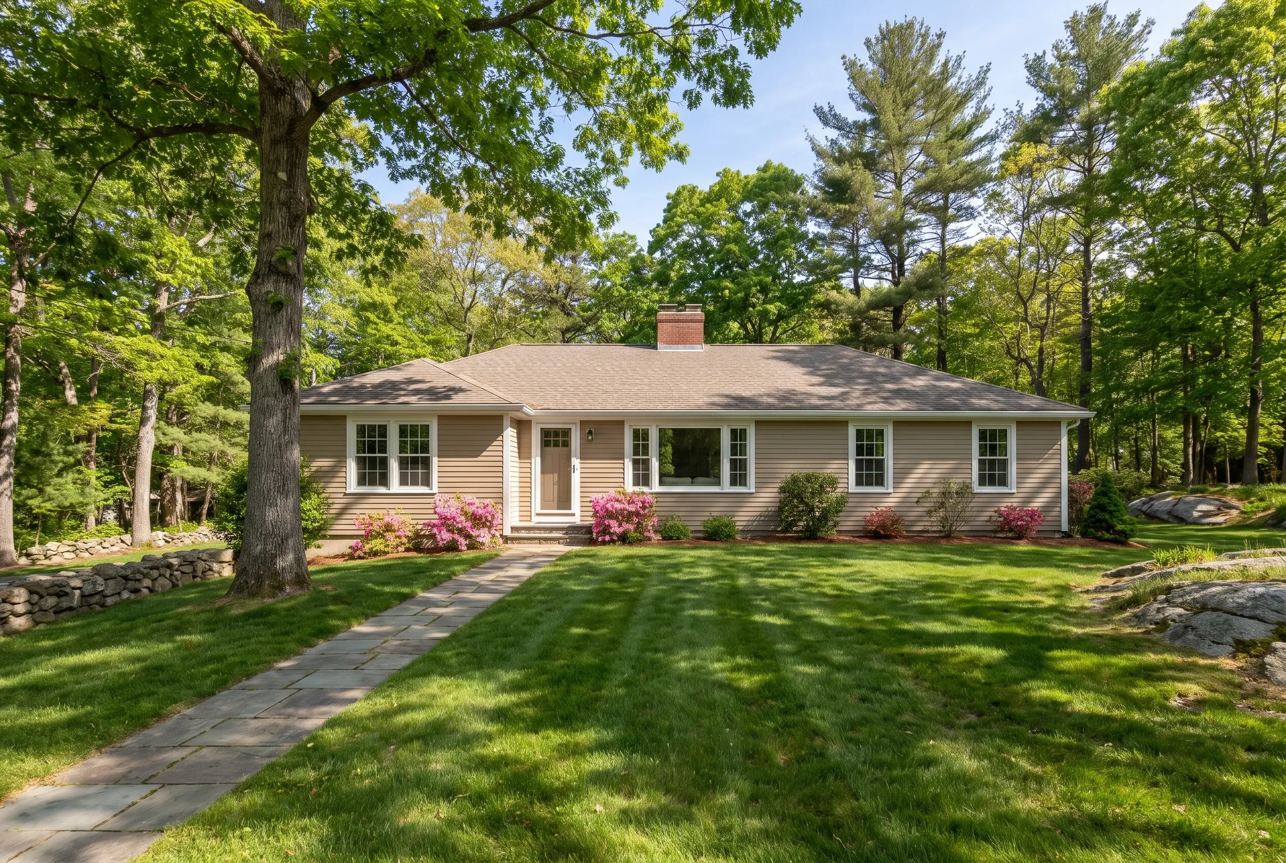 Re-sided ranch in Freeport, ME: Tan vinyl-sided single-story house with white trim and tan roof surrounded by mature trees, blooming pink shrubs, and manicured lawn.