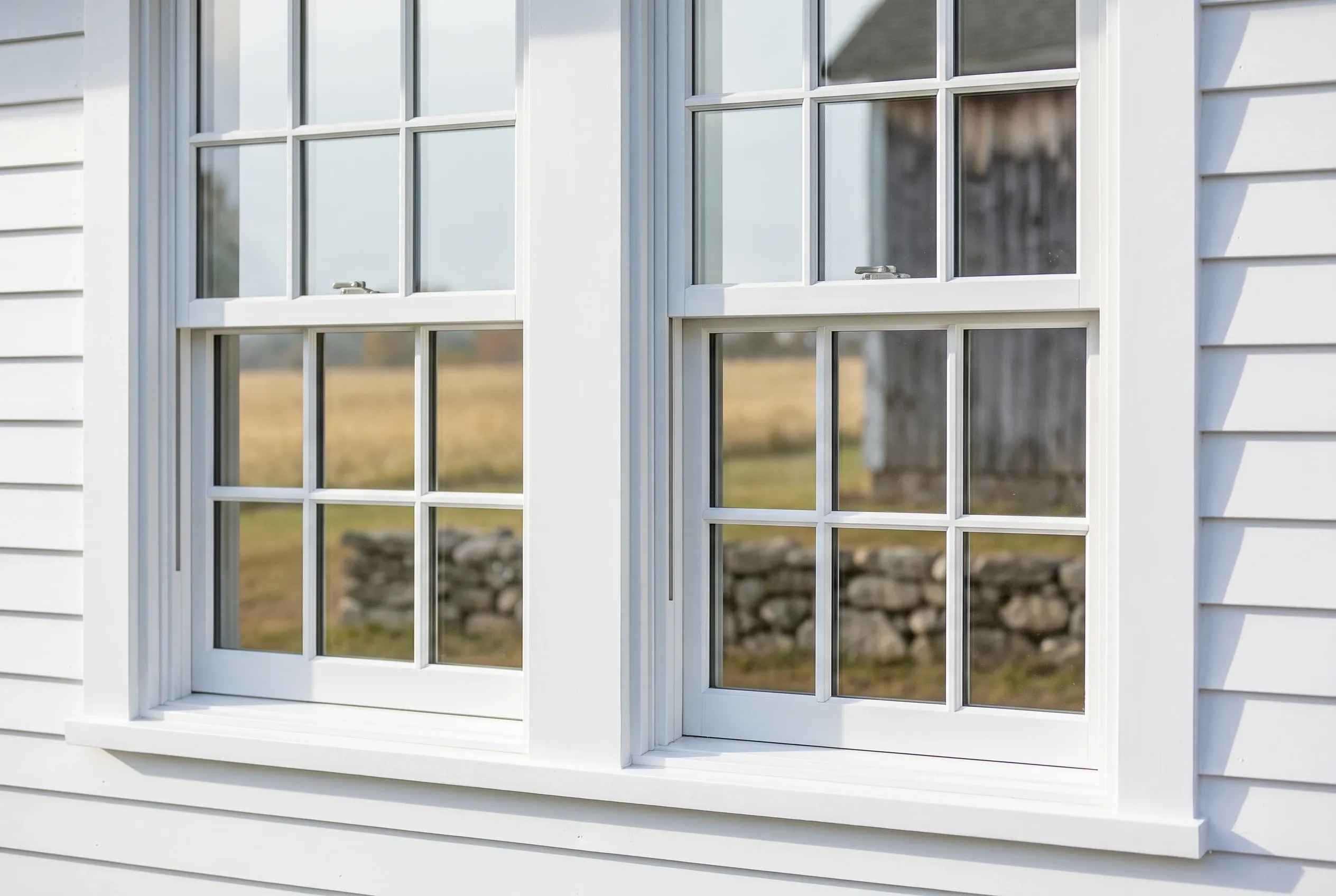 White double-hung windows in Freeport, ME: Two large windows with multiple panes set in white trim on a white-sided residential building with horizontal siding, showing rural landscape through glass.