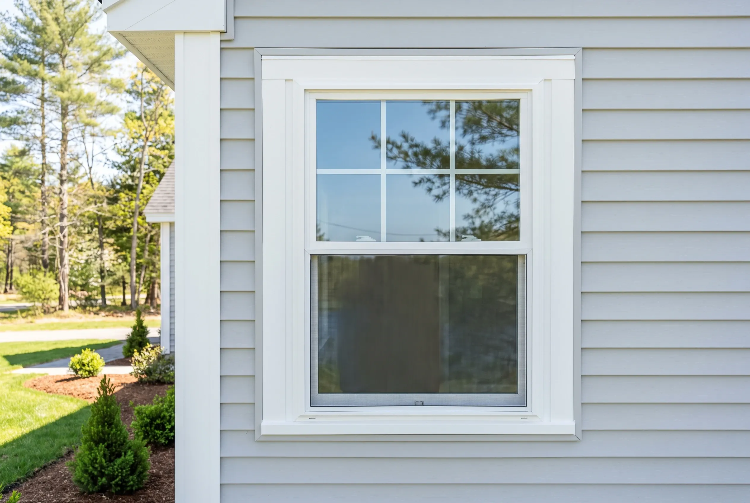 New vinyl windows on Sanford home: Double-hung window with white trim on light gray horizontal siding, evergreen landscaping visible, wooded setting.